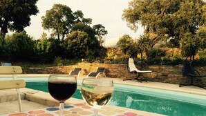 A poolside table with two wine glasses, a stone wall, and lounge chairs in the background.