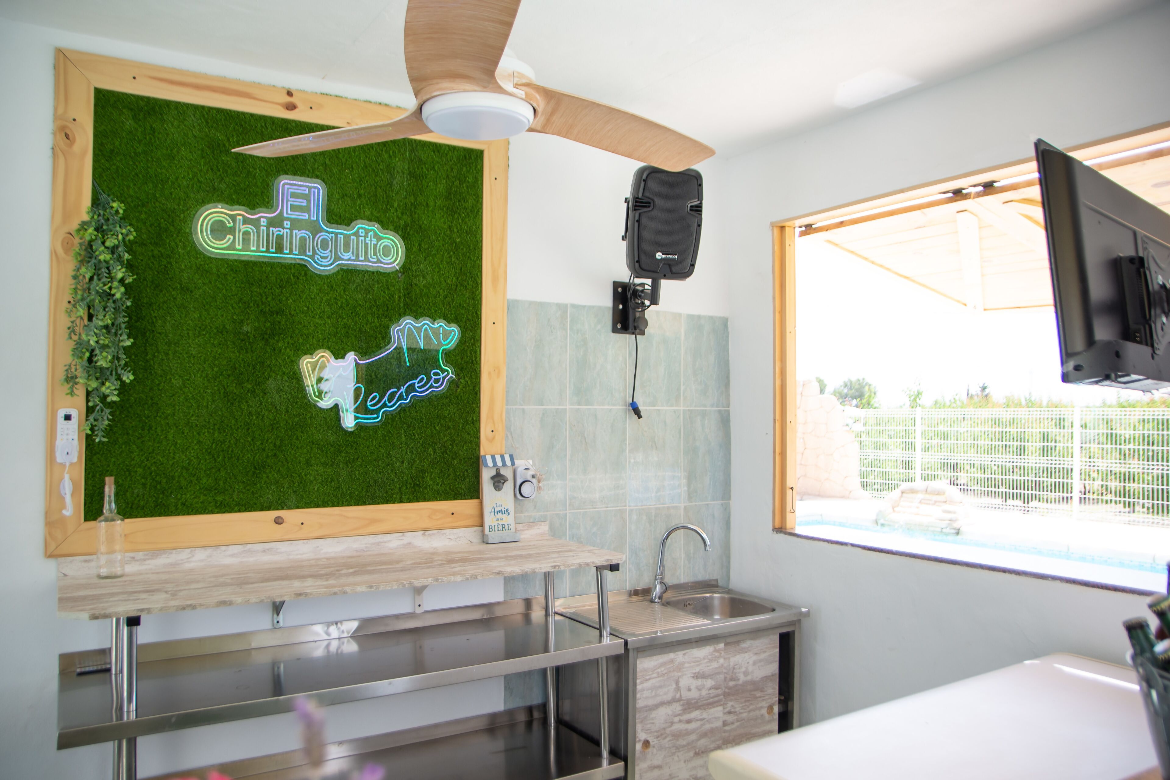 Een keuken met een houten aanrecht, een plafondventilator, een neonbord en een raam met uitzicht op een hek.