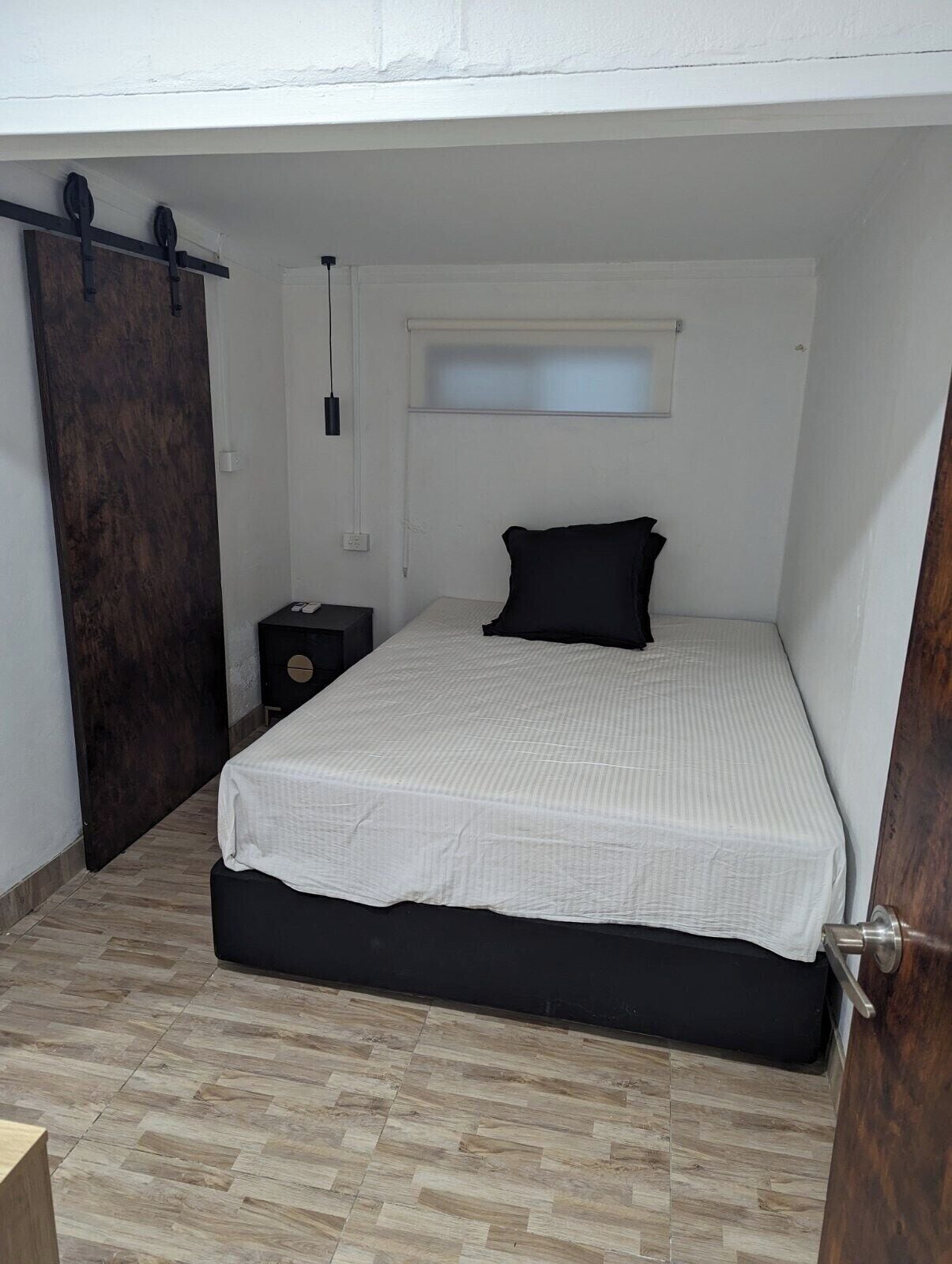 Un dormitori amb un llit, una porta de fusta, una petita taula de nit i una finestra.