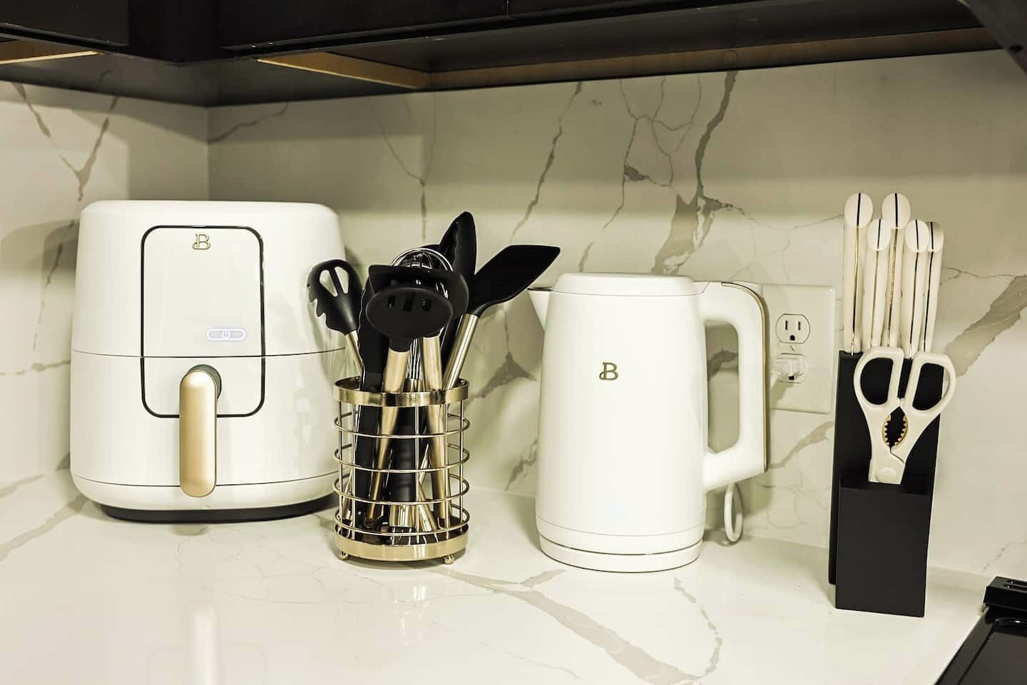 Un plan de travail de cuisine avec un friteuse à air blanche, une machine à café blanche et un porte-ustensiles contenant des ustensiles noirs et dorés.