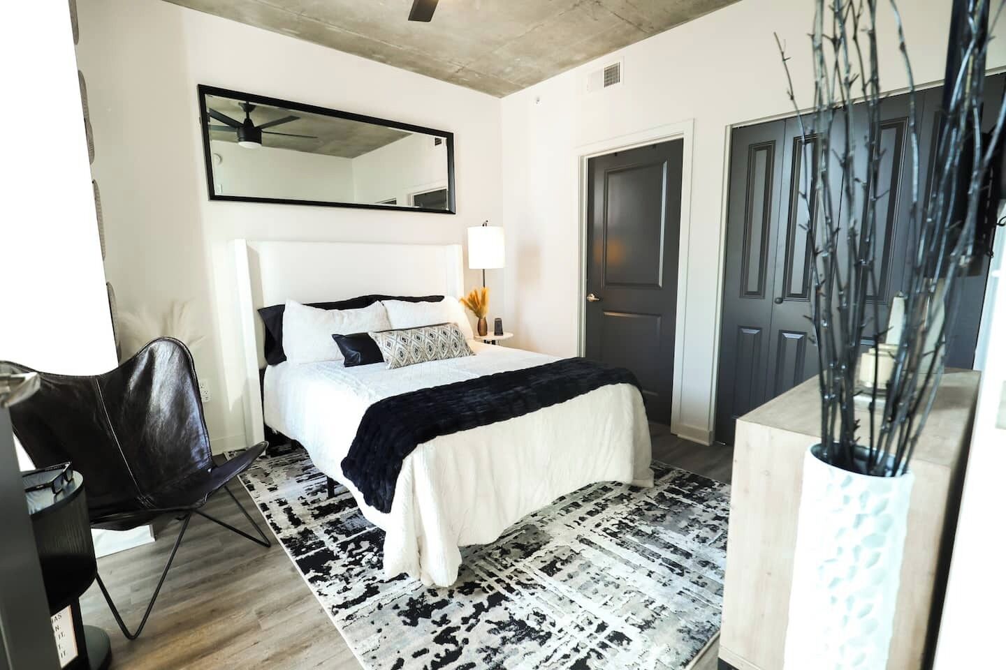 A modern hotel room with a bed, a desk, a chair, a large mirror, and a door.
