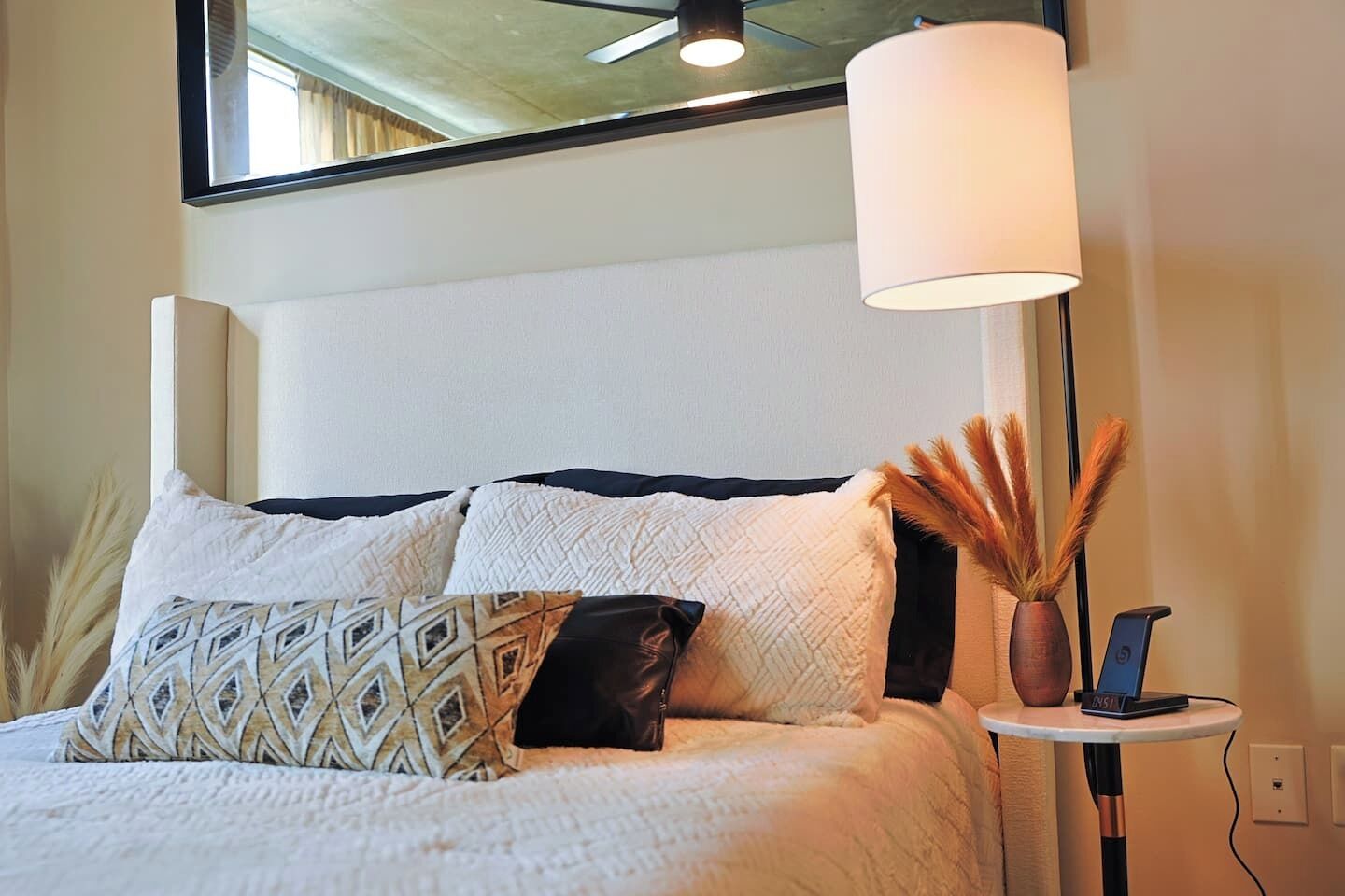 A neatly made bed with patterned pillows, a bedside lamp, and a vase with dried plants on a small table beside the bed.