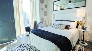 Una habitación de hotel moderna con una cama grande, un espejo redondo, una mesita de noche con una lámpara y vistas a la ciudad a través de la ventana.