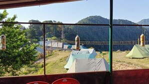 A view from inside a building with a window overlooking a campsite with tents and a rope bridge. - DhaulaPir Adventure (Chamba)
