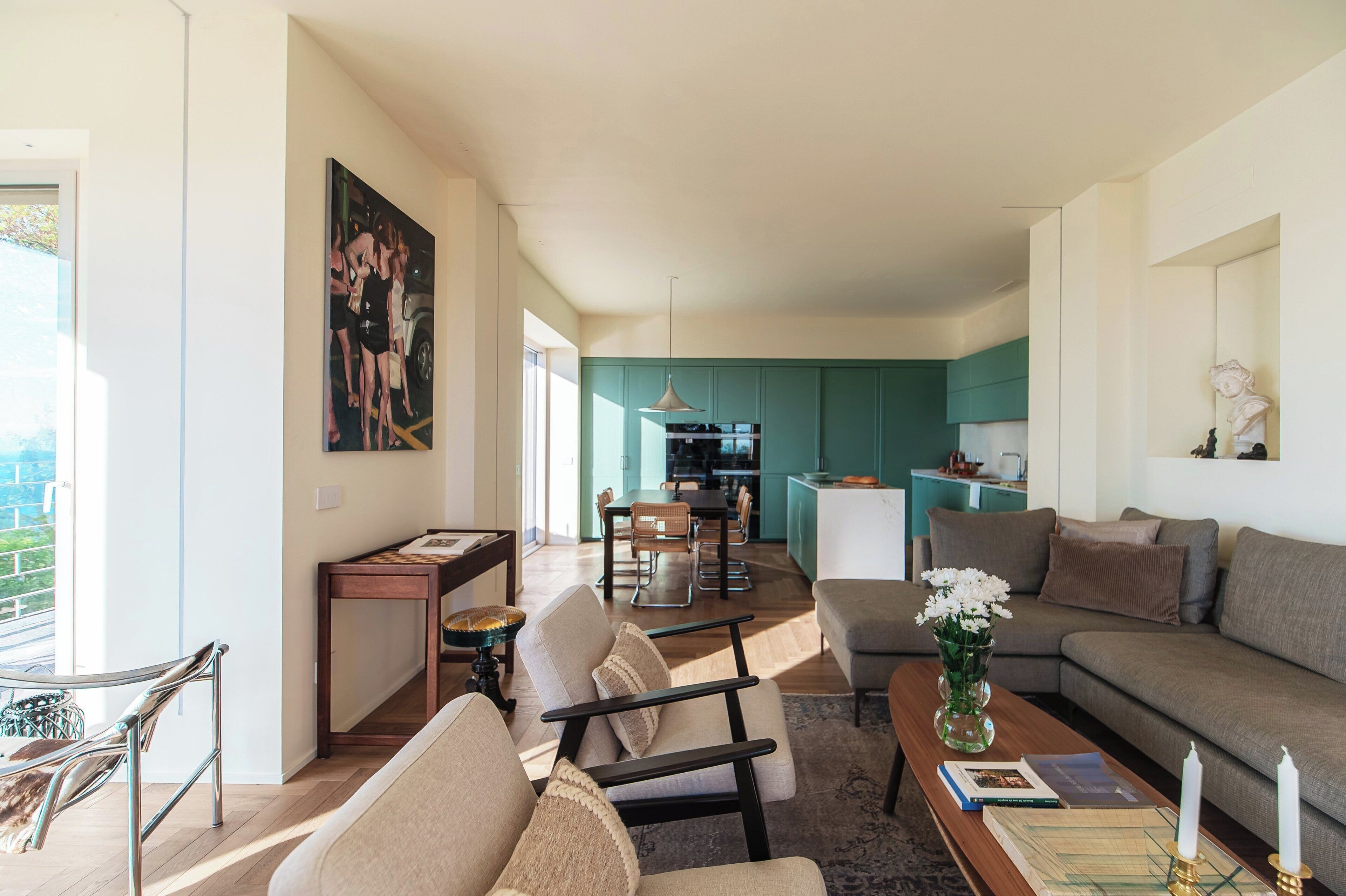 A modern living room with a sofa, chairs, a coffee table, and a dining area with a table and chairs. There is a kitchen in the background with teal cabinets and a hanging light fixture.
