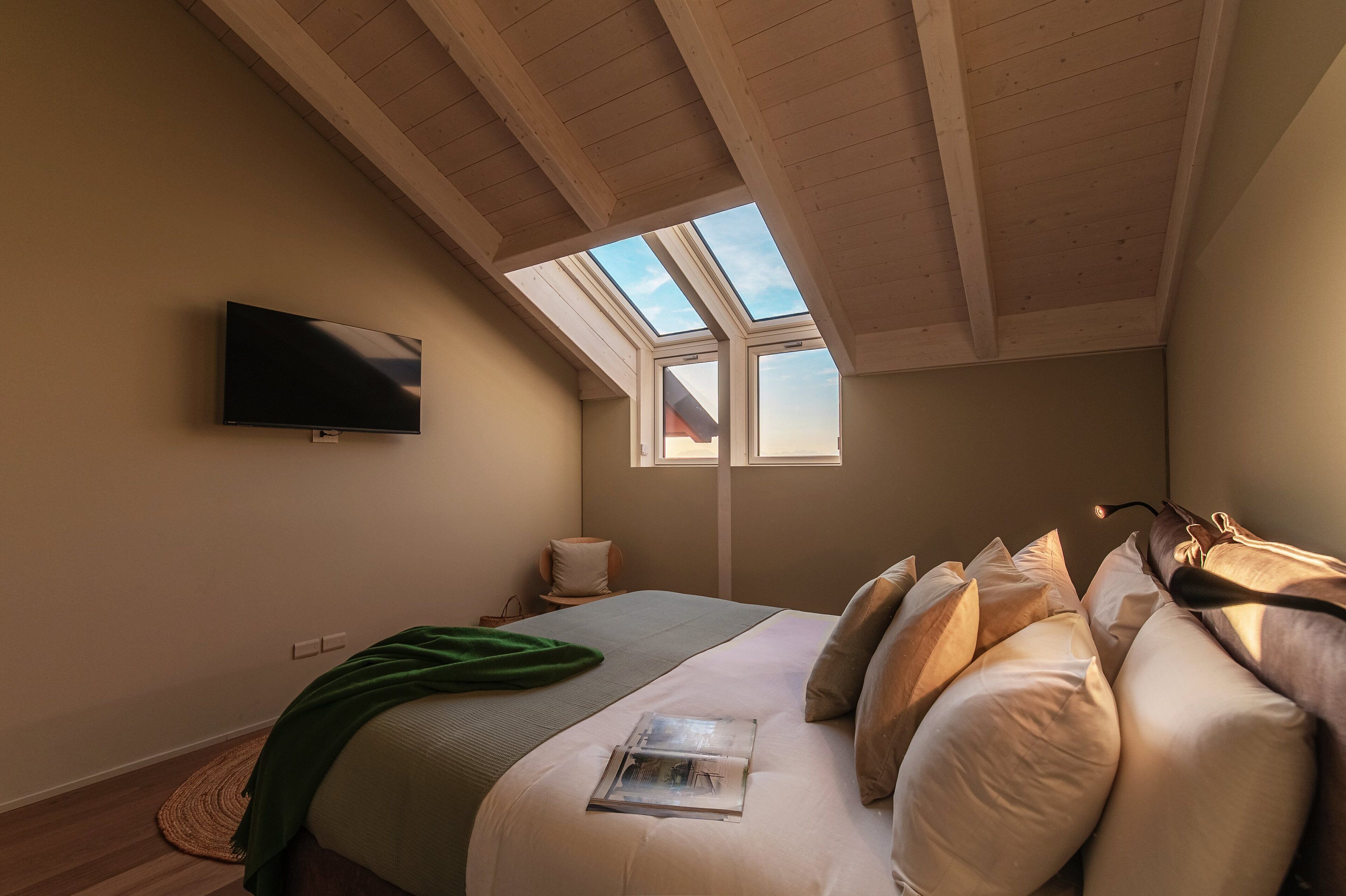 Un dormitorio con techo inclinado, un televisor de pantalla plana montado en la pared, una cama con varias almohadas, una mesita de noche con lámpara y un ventanal que deja entrar luz natural.