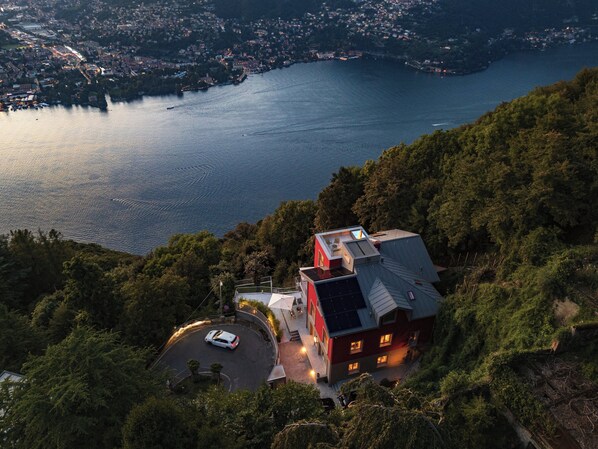 A high vantage point view of a coastal town with a prominent red building featuring a rooftop terrace, surrounded by lush greenery and overlooking a calm sea. - Villa Rossa (Brunate)