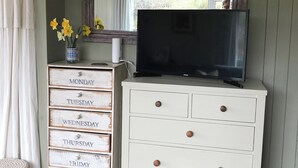 Une pièce avec une commode blanche dotée de tiroirs étiquetés avec les jours de la semaine, un téléviseur à écran plat posé sur le dessus, un miroir au-dessus du téléviseur et une fenêtre avec des rideaux.