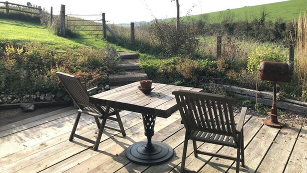 Une terrasse en bois avec une table et deux chaises, entourée d’un paysage rural agrémenté d’une clôture et de verdure.
