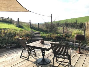 A wooden deck with a table and two chairs, surrounded by a rural landscape with a fence and greenery.