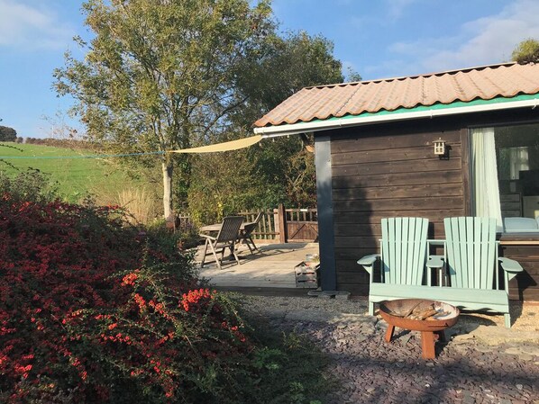 A wooden cabin with a tiled roof, a porch with green Adirondack chairs, and a fire pit.