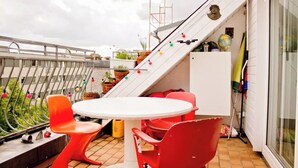 A rooftop terrace with a white table and red chairs, potted plants, and a staircase leading to an upper level. - From November 4-6 months 80sqm apartment with roof terrace in Neukölln (Berlin)