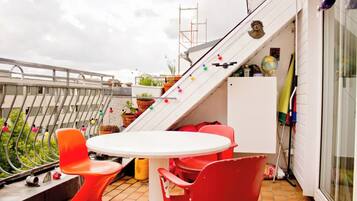 Eine Dachterrasse mit weißem Tisch und roten Stühlen, Blumentöpfen und einer Treppe, die zu einer oberen Ebene führt.