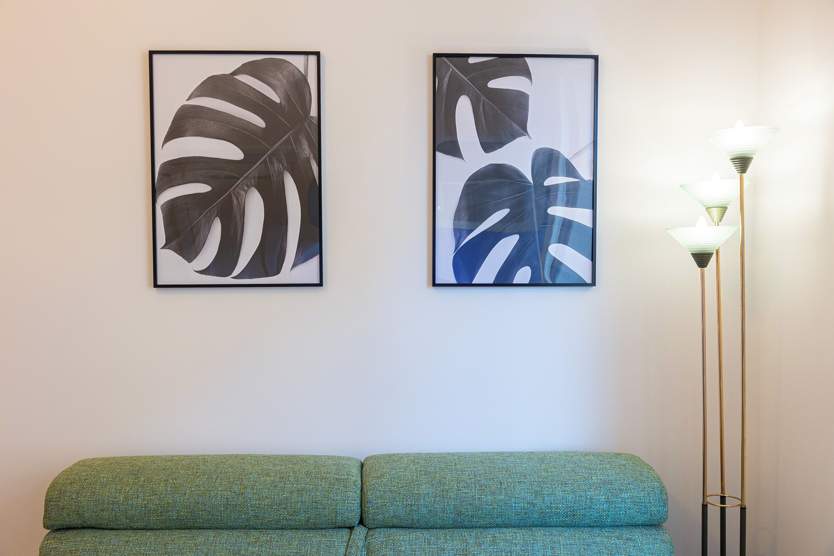 Una habitació amb un sofà verd, dos quadres amb fulles tropicals al mur, i una làmpada de peu alta amb múltiples fonts de llum.