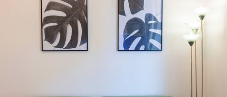 A room with a green sofa, two framed tropical leaf artworks on the wall, and a tall floor lamp with multiple light sources.