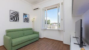 Egy szoba, melyben egy zöld kanapé, egy televízió, egy ablak, ahonnan kilátás nyílik az épületekre, és két, keretbe foglalt műalkotás található a falon.