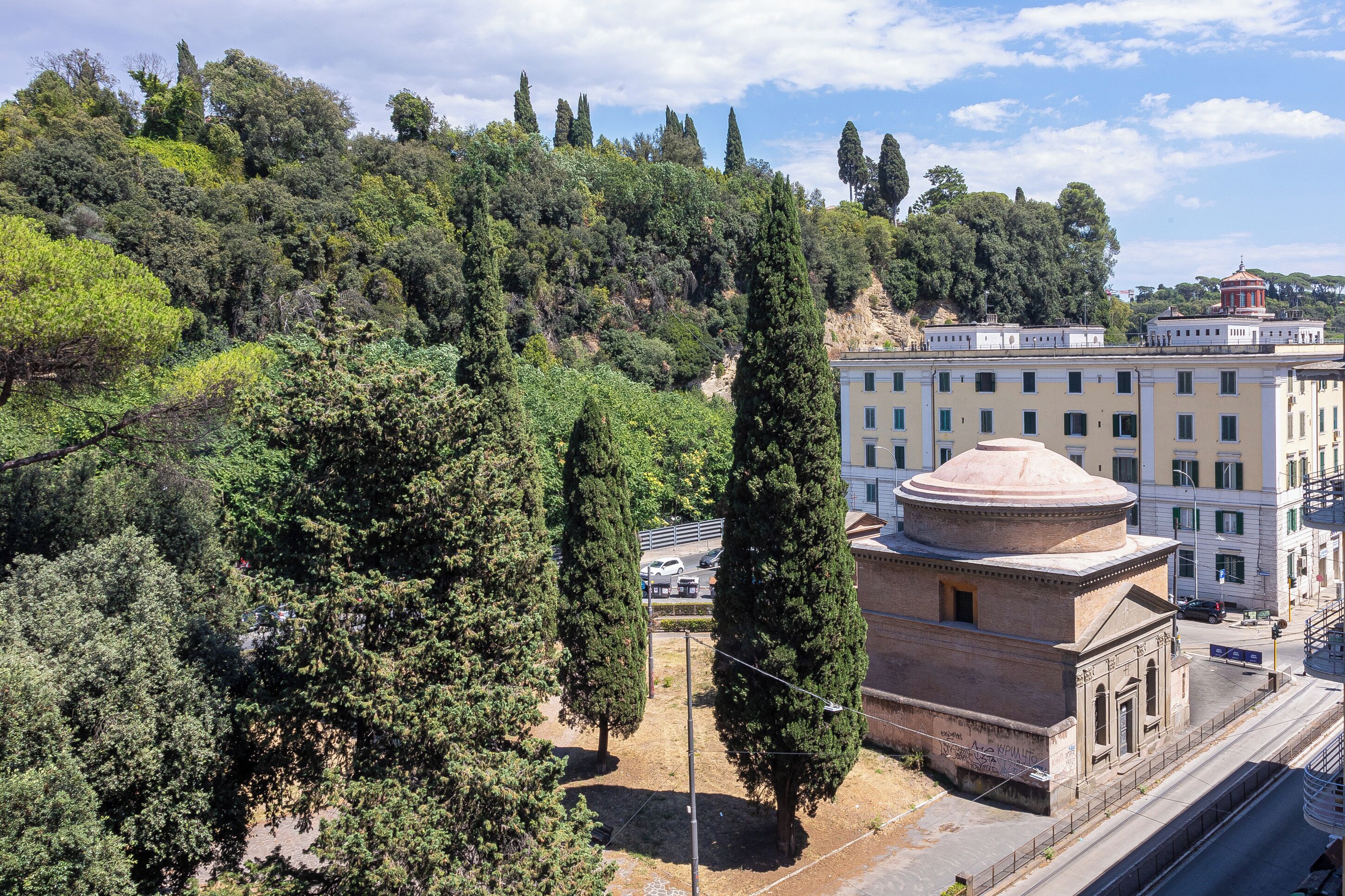 A building with a dome structure, surrounded by trees and a hilly landscape.