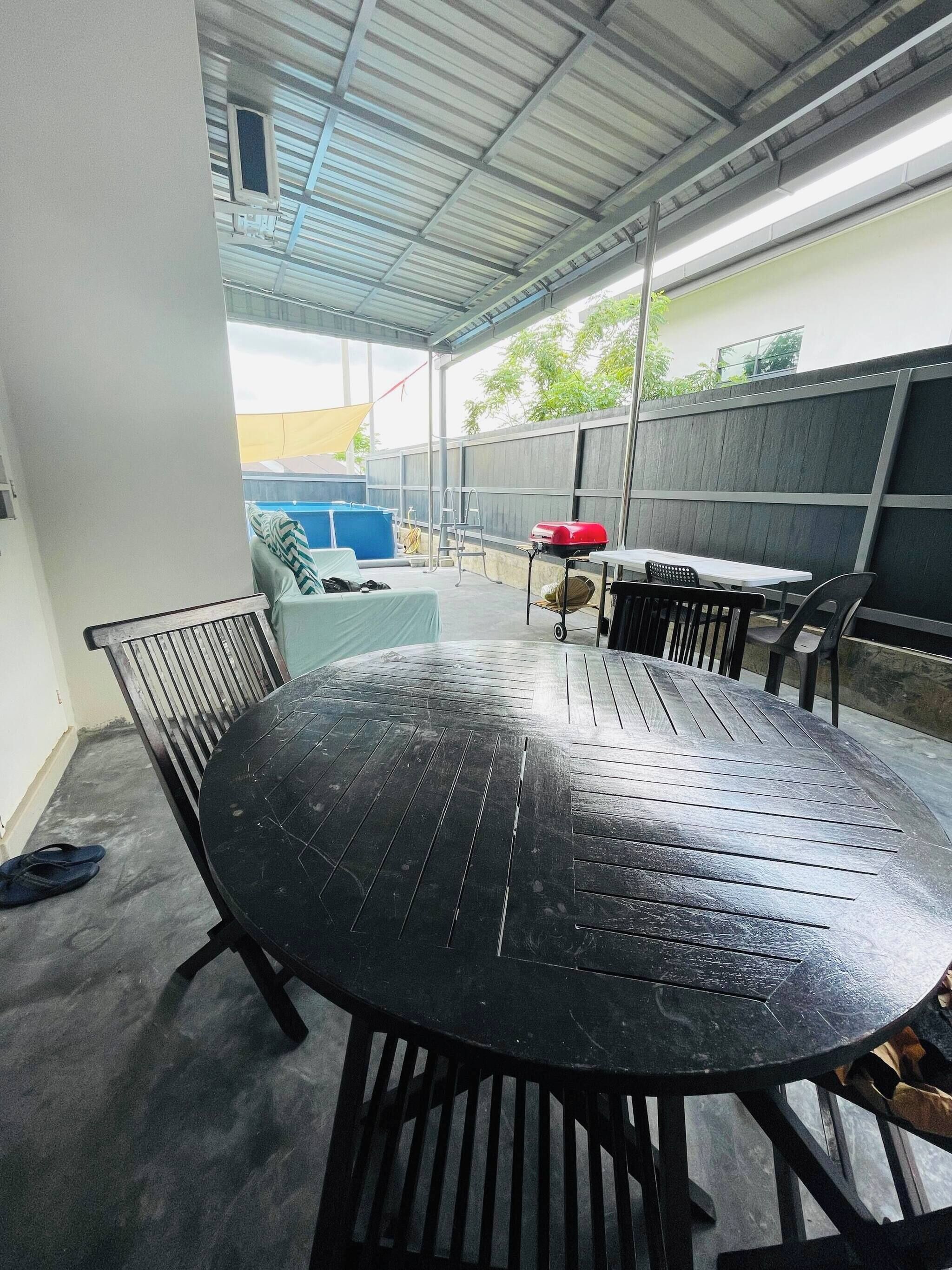Um alpendre coberto com mesa e cadeiras redondas, churrasqueira e piscina azul ao fundo.