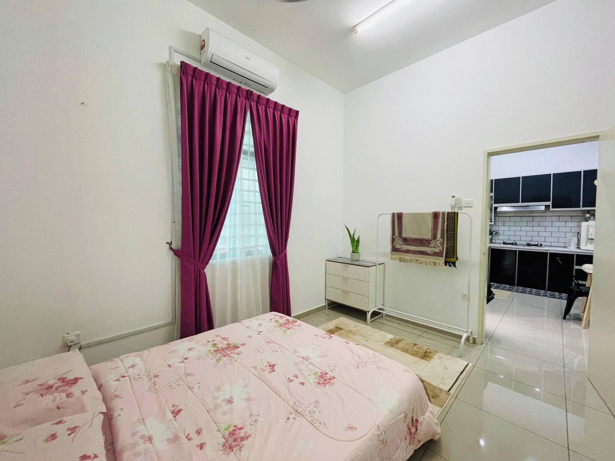 Quarto com cama, janela com cortinas roxas, mesa de cabeceira com planta e vista para uma cozinha.