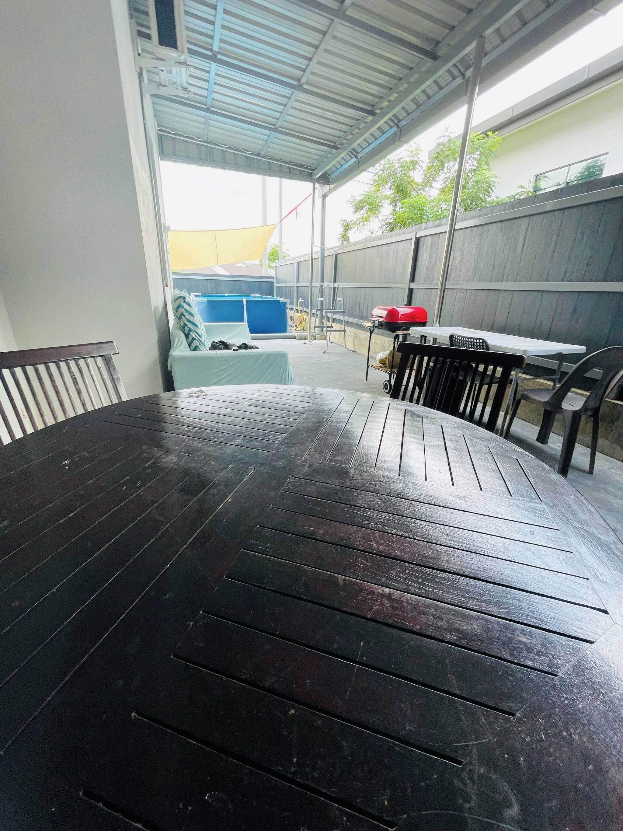 Ein Außenbereich mit einer Terrasse, einem runden Holztisch und Stühlen, einem überdachten Sitzbereich und einem Pool im Hintergrund.