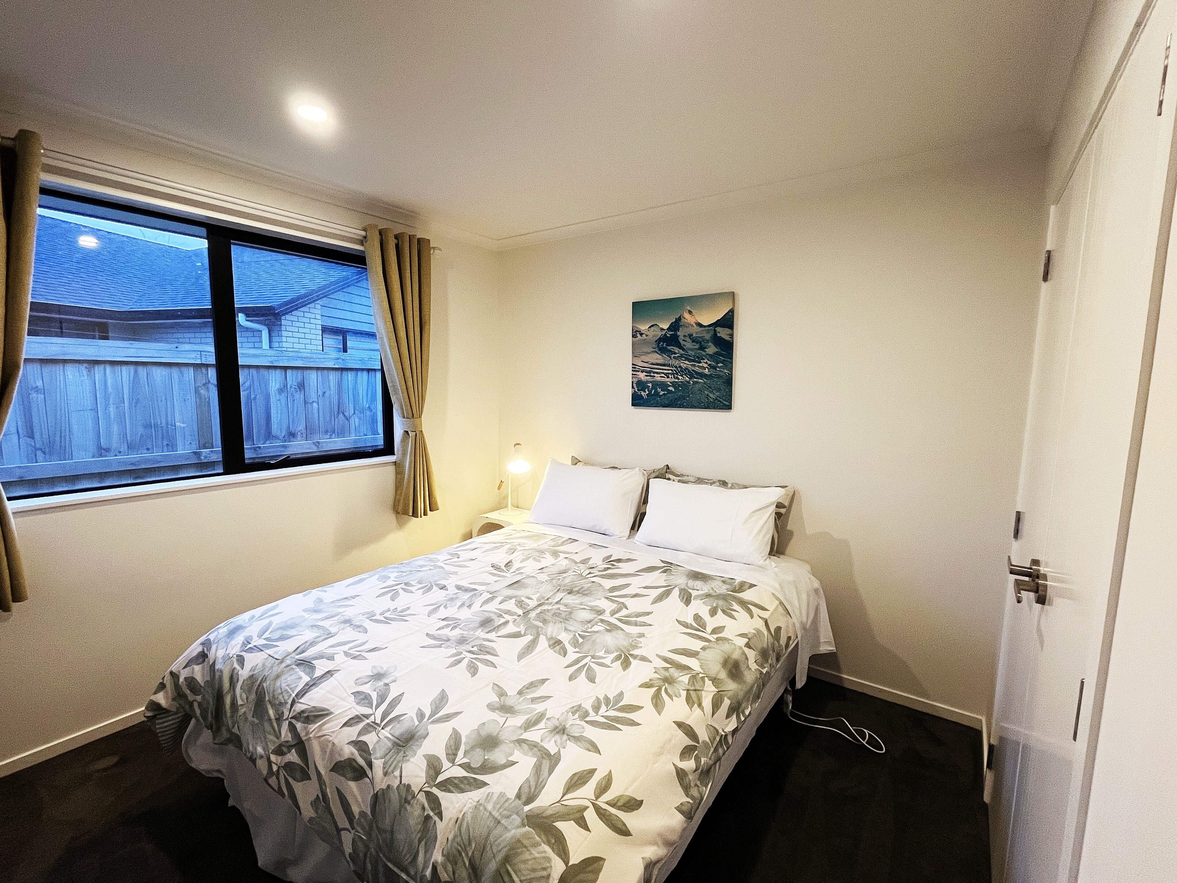 Habitación de hotel con cama, ventana con cortinas, mesita de noche con lámpara y un cuadro colgado en la pared.