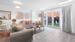Ein modernes Wohnzimmer mit Sofa, Sessel und Couchtisch. Es gibt einen Essbereich mit Tisch und Stühlen. Das Zimmer verfügt über eine große Schiebeglasdoor, die zu einem Garten führt.