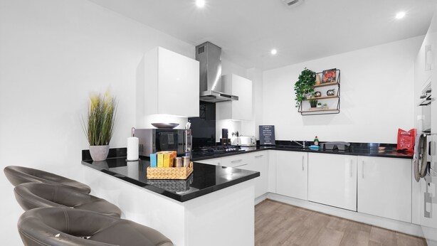 A modern kitchen with white cabinets, a black countertop, and a built-in oven. There are bar stools and a potted plant on the counter. - Spacious 6 Bedroom House in Denton Way (Slough)