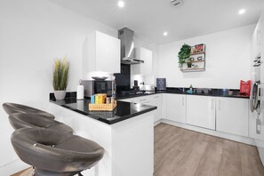 A modern kitchen with white cabinets, a black countertop, and a built-in oven. There are bar stools and a potted plant on the counter. - Spacious 6 Bedroom House in Denton Way (Slough)
