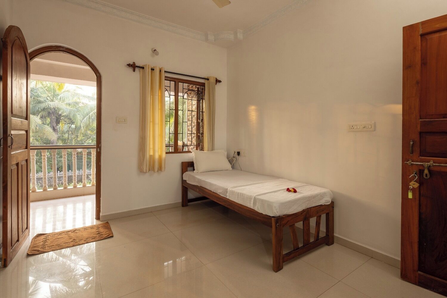 Una habitación pequeña y limpia con una cama individual, una ventana con cortinas, una puerta y un balcón con vistas a palmeras.