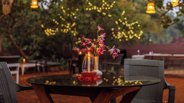 A table with a vase of flowers and scattered coins, surrounded by chairs and string lights in an outdoor setting. - Cashew Garden Homestay (Agonda)