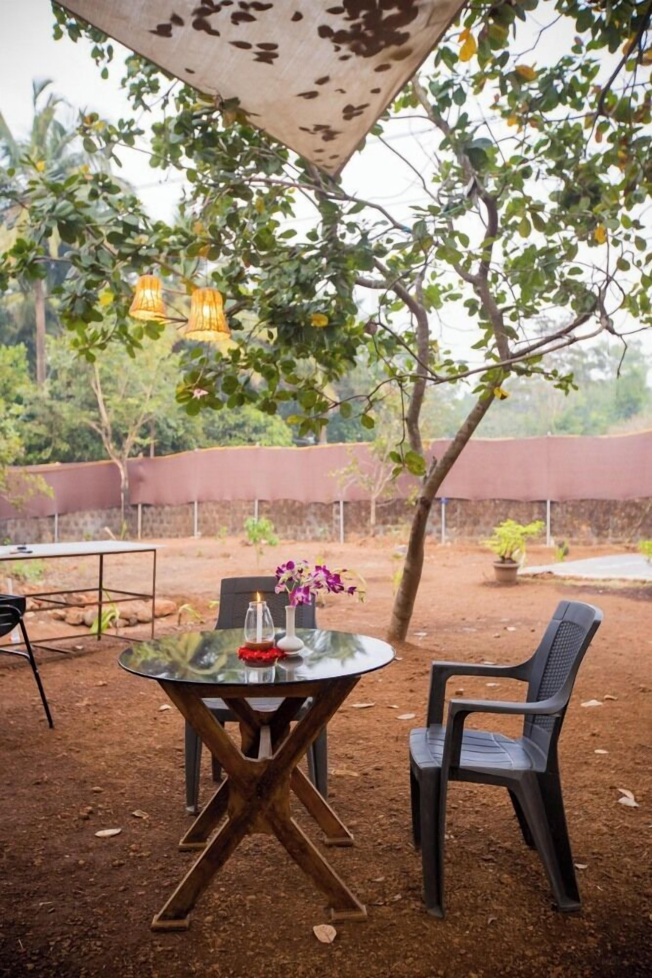 An outdoor dining area with a round table and two chairs, surrounded by trees and a covered seating area.