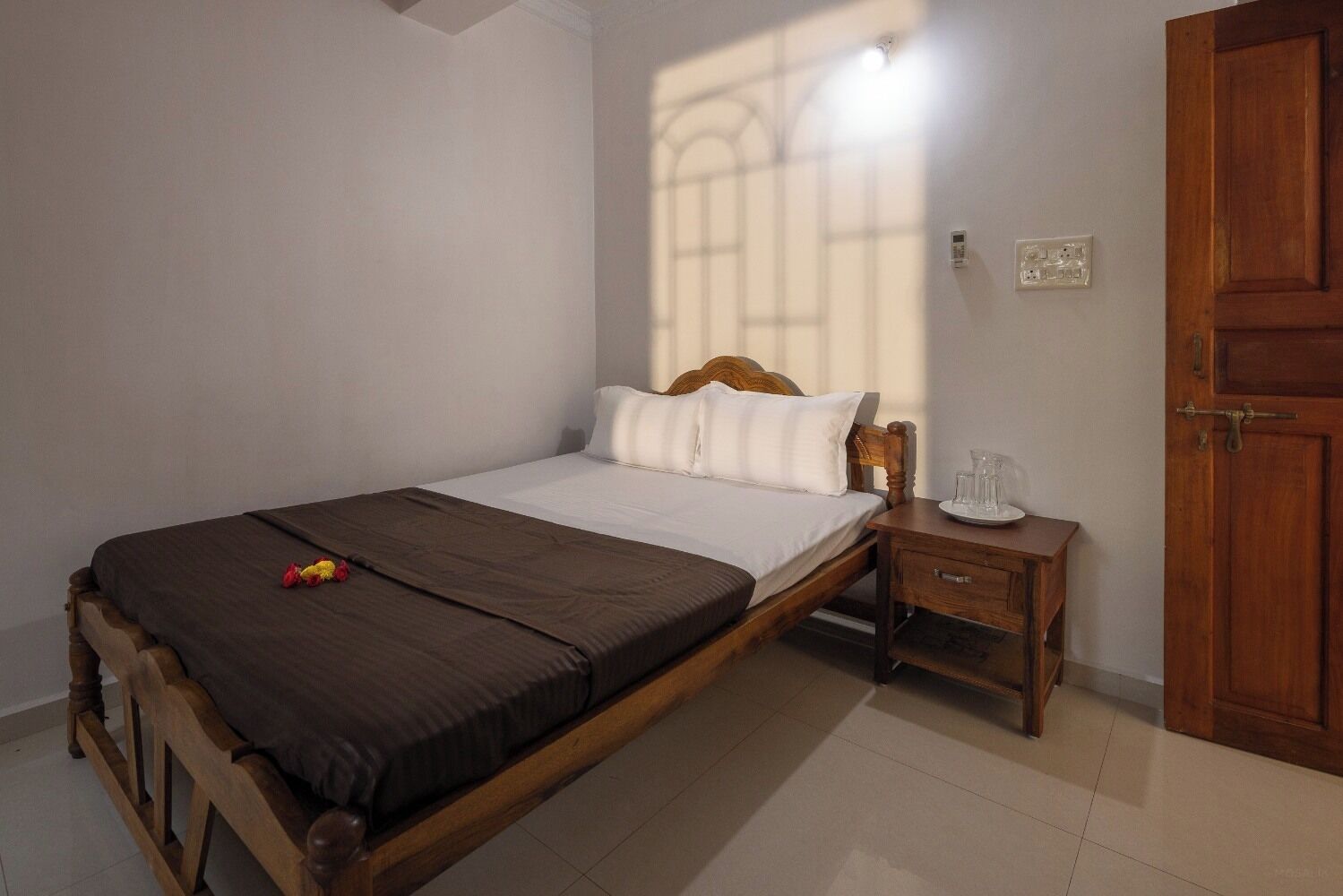 A neatly made bed with a brown cover, white pillows, and a wooden headboard. There is a small wooden nightstand with a glass of water and a plate beside the bed. A wooden door is visible to the right.