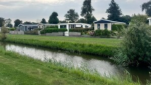 A row of modern mobile homes with a grassy area and a canal in the foreground. - Cozy chalet near the sea. Pet friendly! (Schagen)