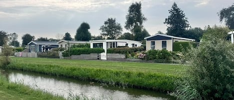 A row of modern mobile homes with a grassy area and a canal in the foreground.