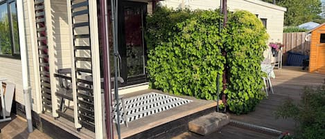 En overdækket patio med træterrasse, en metalpergola og en frodig grøn hæk.