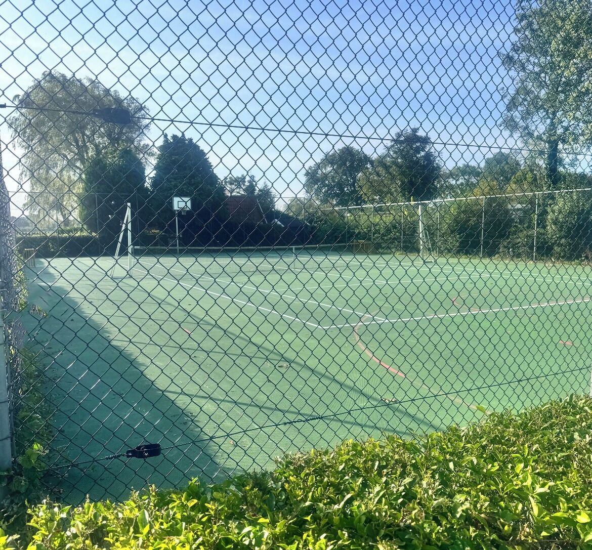 Een tennisbaan omheind door een hek van gaas met een basketbalveld op de achtergrond.