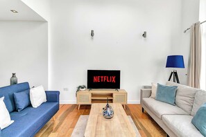 Una sala de estar con un televisor de pantalla plana mostrando 'NETFLIX', un sofá azul, un sofá gris, una mesa de centro de madera y una lámpara.