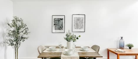 A dining area with a wooden table set for four, two framed artworks on the wall, and a potted plant on the left.