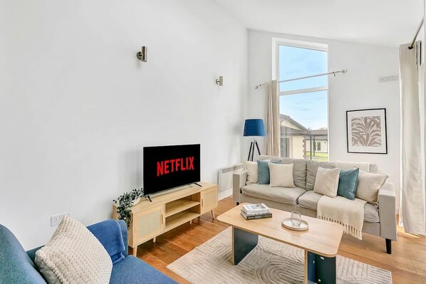 Una sala de estar moderna con un televisor mostrando 'NETFLIX', un sofá, una mesa de centro y una ventana con cortinas.