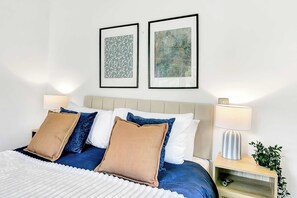 A neatly made bed with a white headboard, blue and beige pillows, and a white comforter. There are two framed abstract paintings above the bed. - Bright & Modern 2 Bed Holiday Chalet nr Sidmouth (Sidmouth)