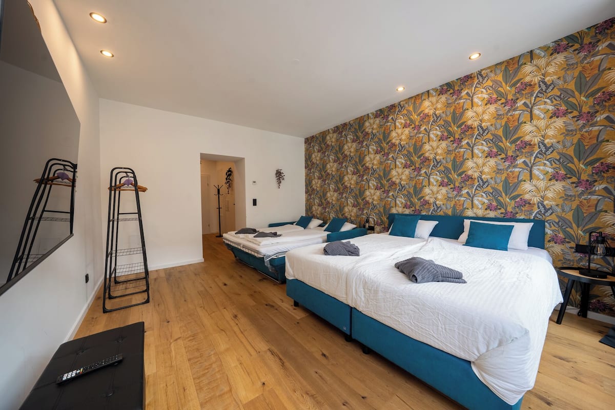 A hotel room with two beds, a wooden floor, a floral wallpaper, and a black TV stand.