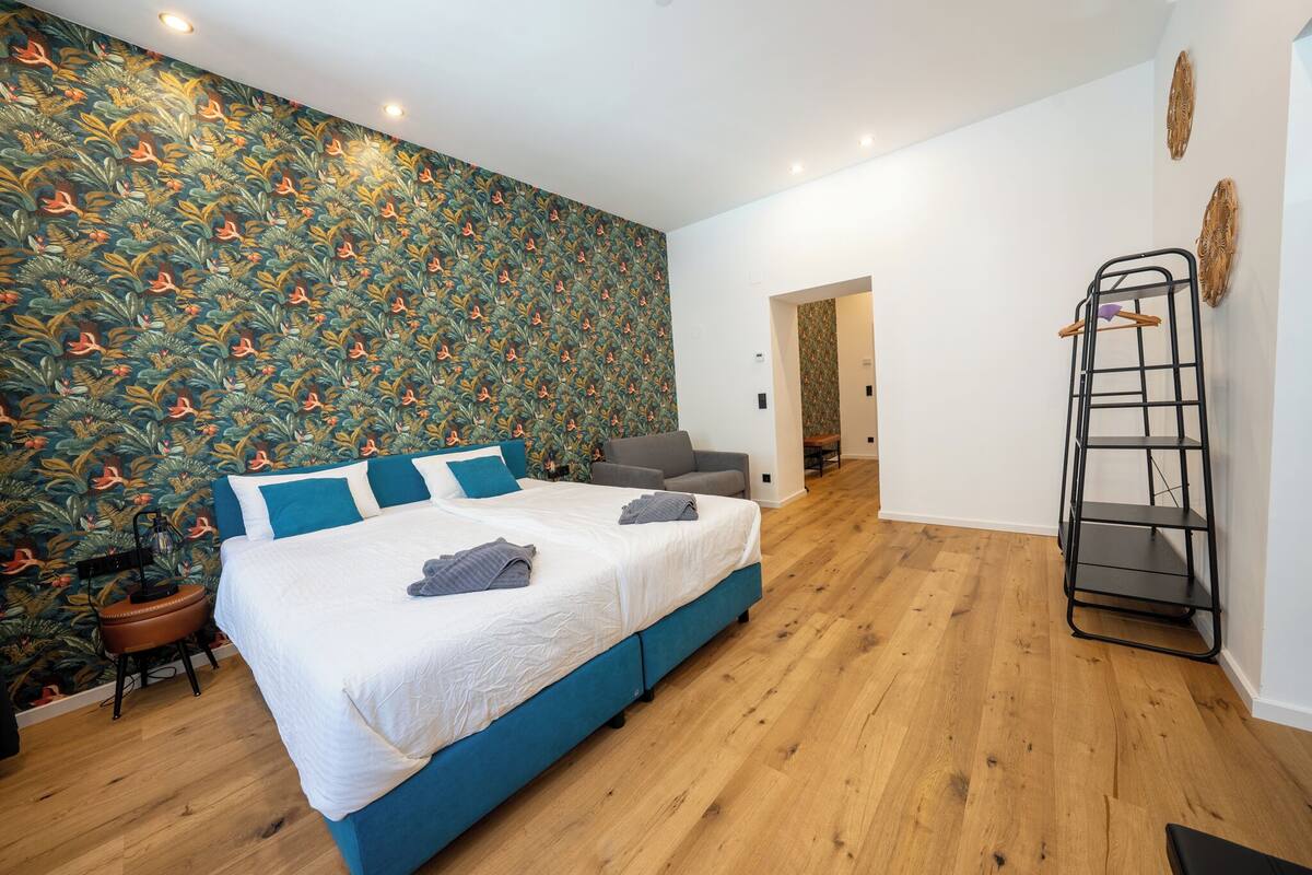 A modern bedroom with a large bed, a grey sofa, a wooden floor, and a decorative wall.