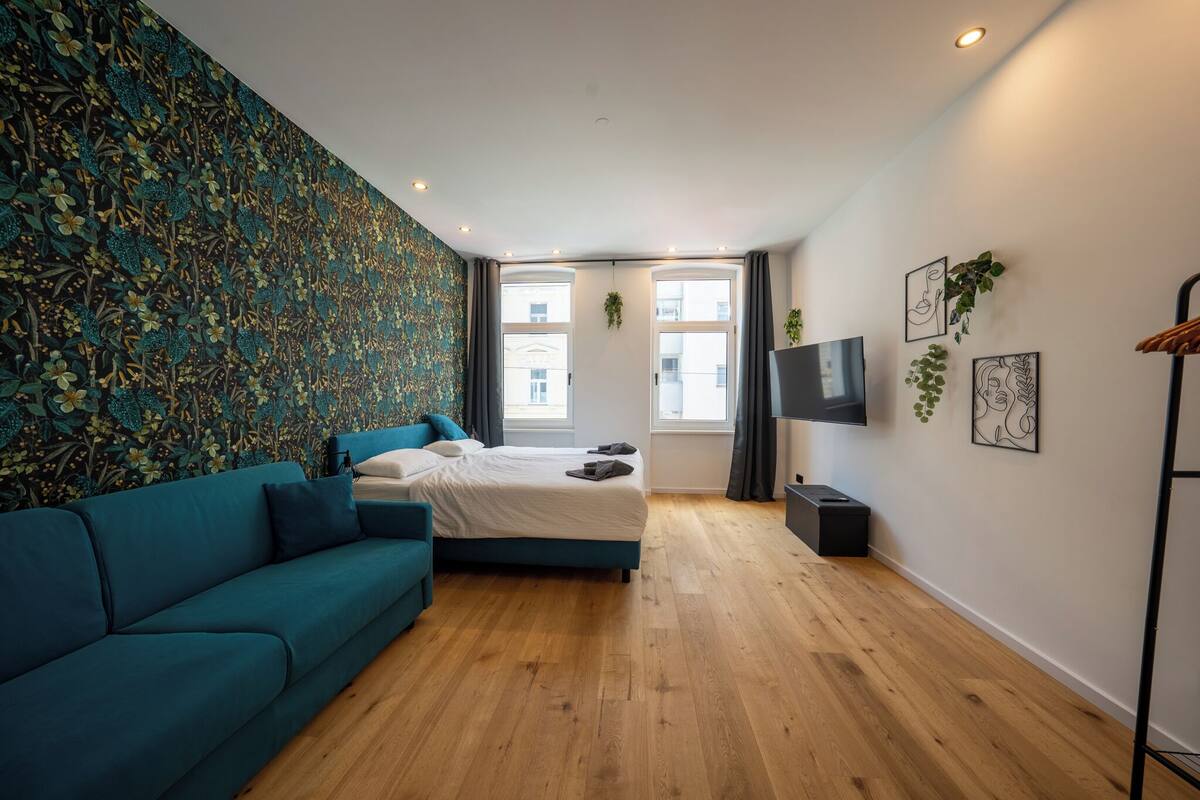 A modern bedroom with a teal sofa, a bed, a TV, and a large window with a view of buildings.