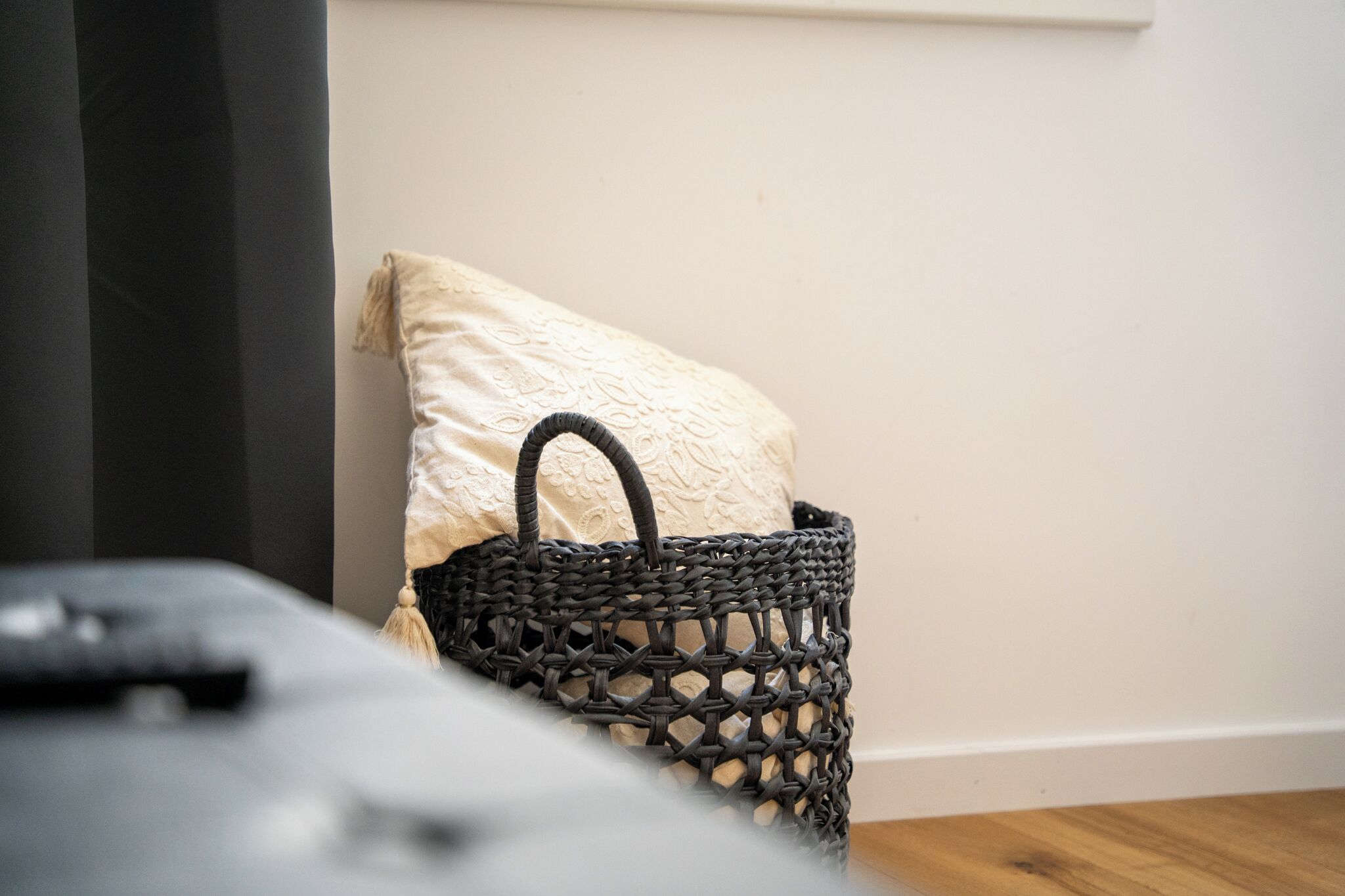 A wicker basket with a handle and a cushion on a wooden floor, next to a dark-colored sofa.