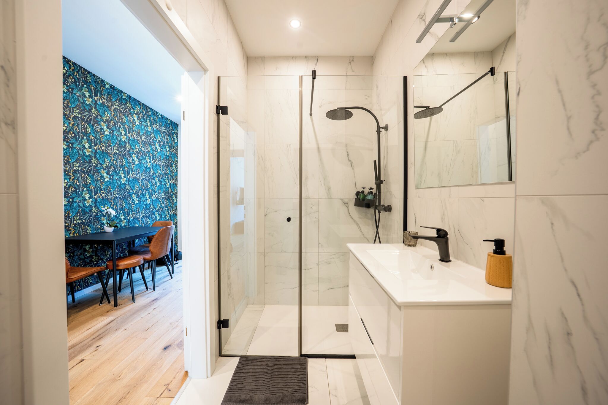 A modern bathroom with a glass-enclosed shower, white marble walls, and a white sink with a soap dispenser.
