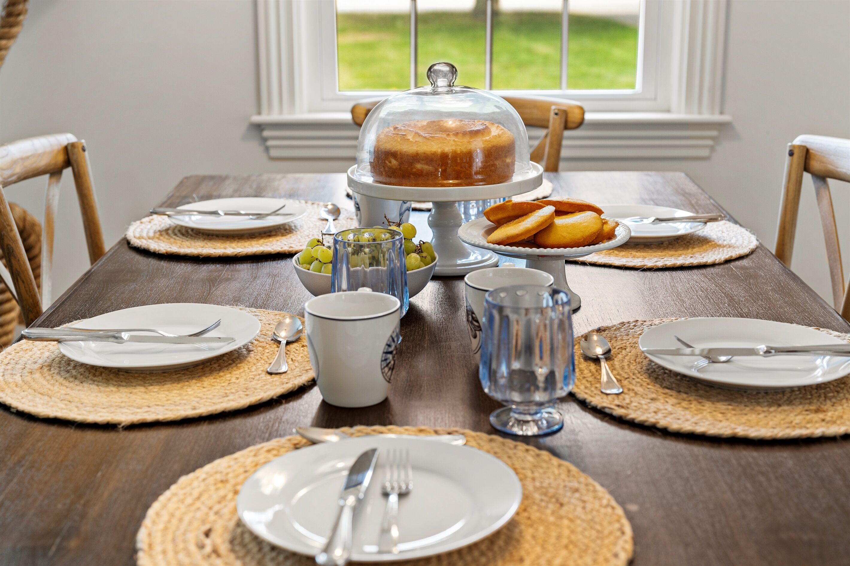Een eettafel gedekt met borden, bestek, een glazen karaf, een schaal druiven en een taart onder een deksel, alles op een geweven placemat.