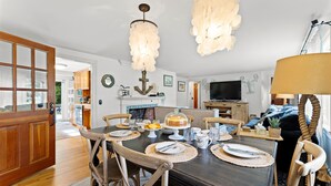 A dining area with a dark wooden table set for four, surrounded by wooden chairs. A chandelier hangs above, and a television is visible in the background.