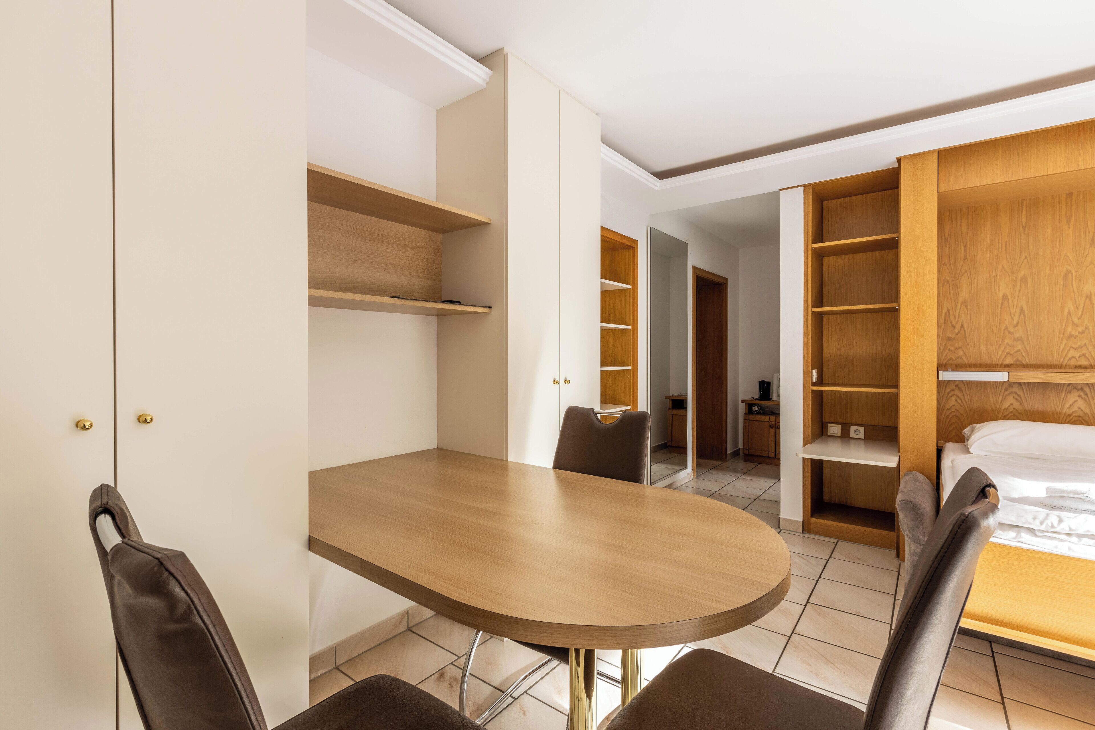 A hotel room with a wooden dining table, two chairs, a bed with a wooden headboard, and built-in shelving.