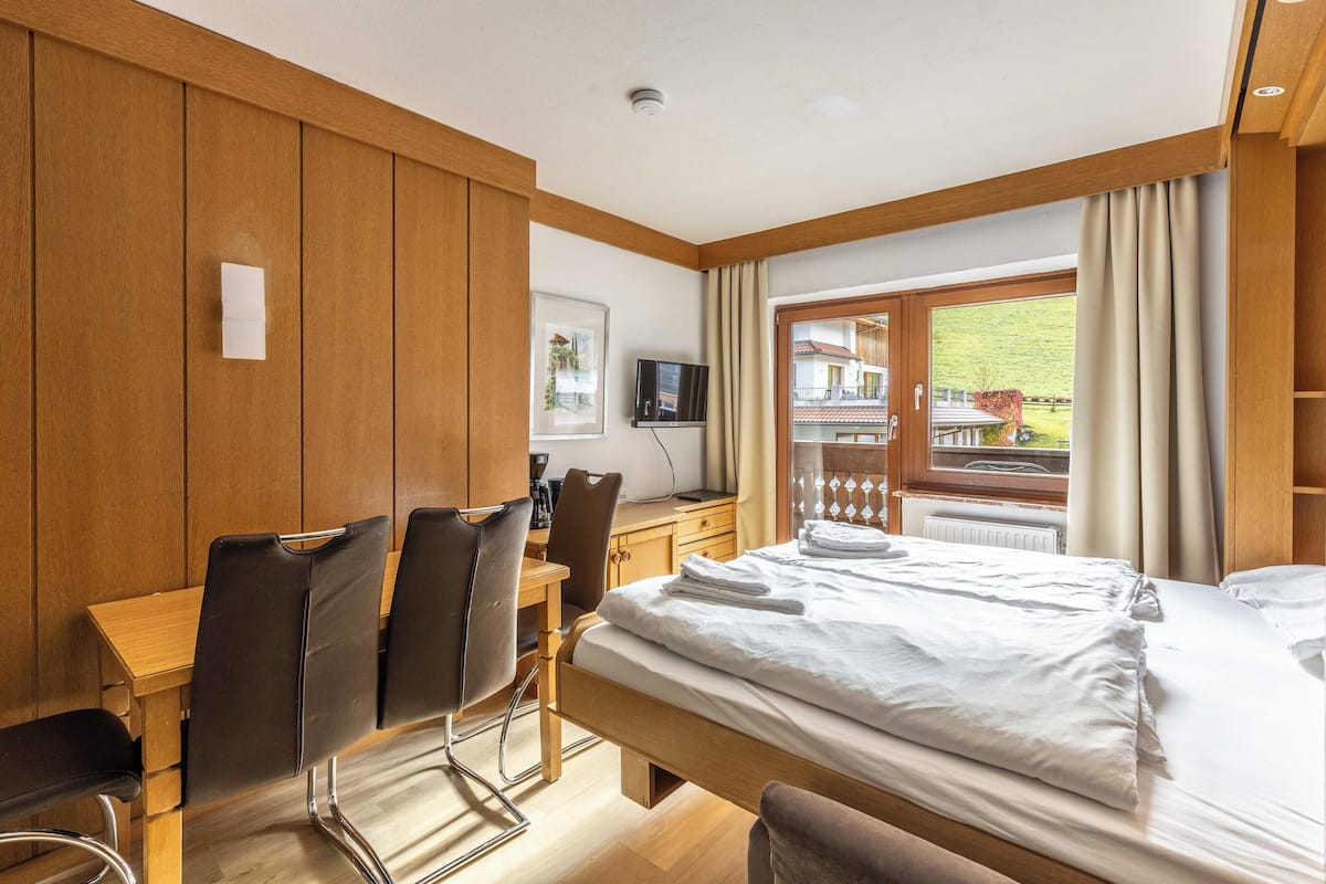 Habitación de hotel con pared de paneles de madera, cama con ropa blanca, escritorio con silla, televisor y ventana con cortinas.