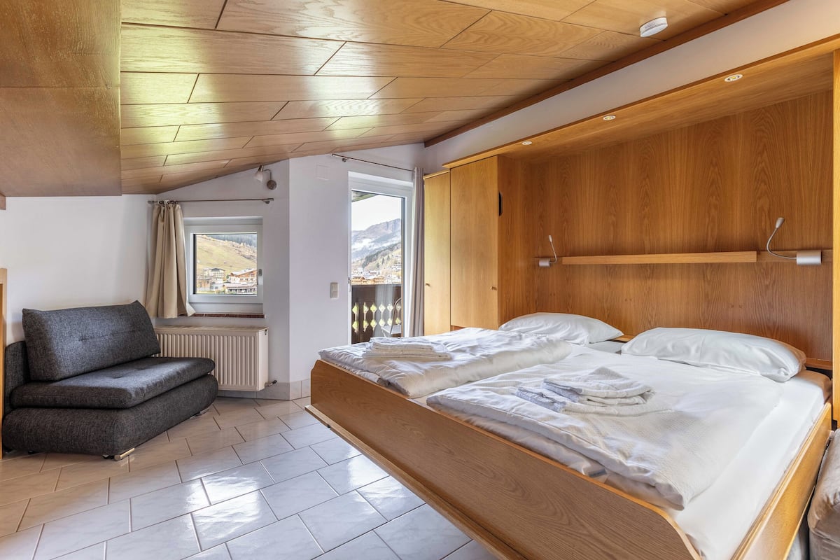 Un dormitorio moderno con una cama de madera, un sofá gris y una ventana con vistas a montañas.